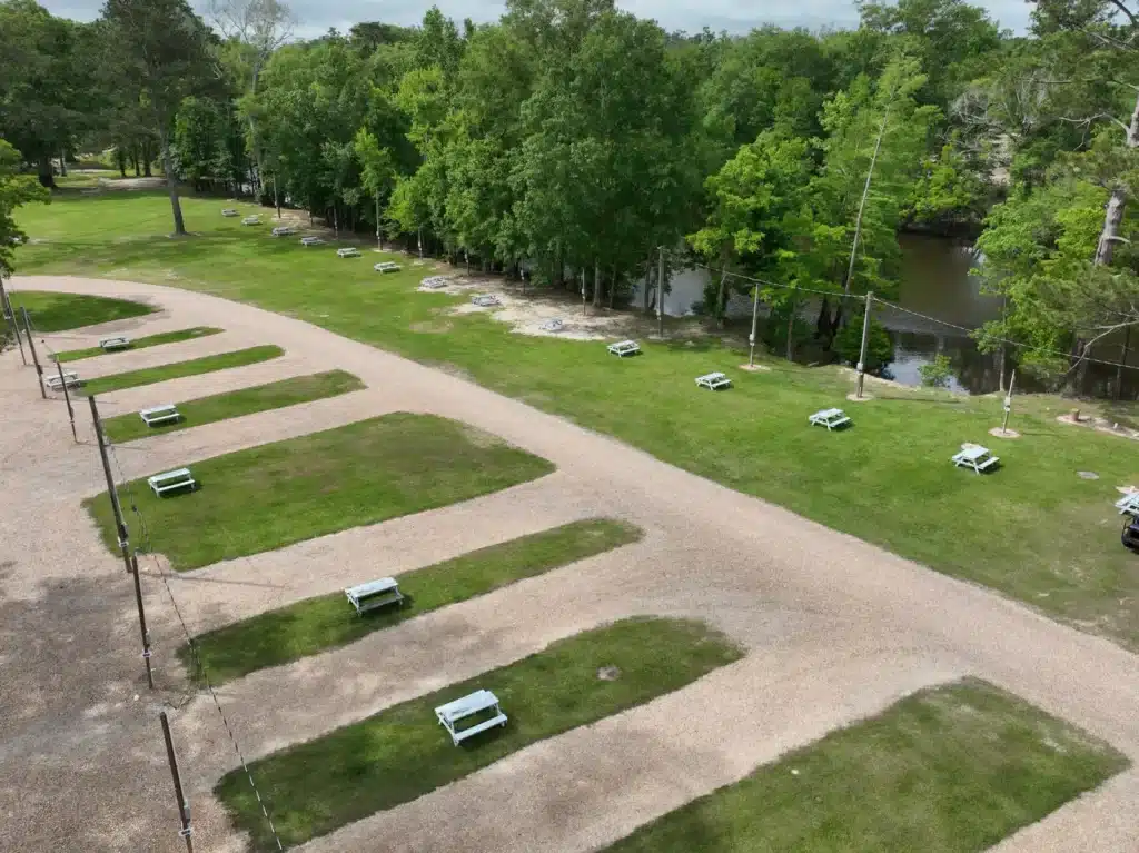 Aerial view of empty gravel RV sites with white picnic tables on grass strips beside a wooded river.