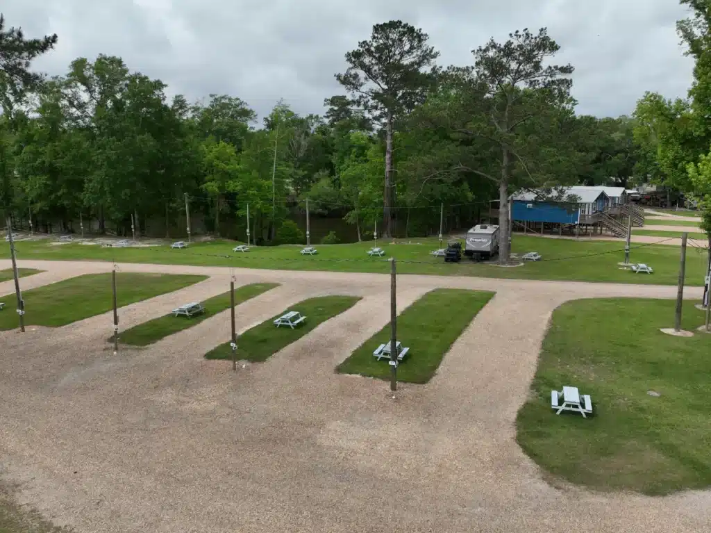 Aerial view of gravel RV park sites with grassy strips, white picnic tables, utility poles, a travel trailer, and pond