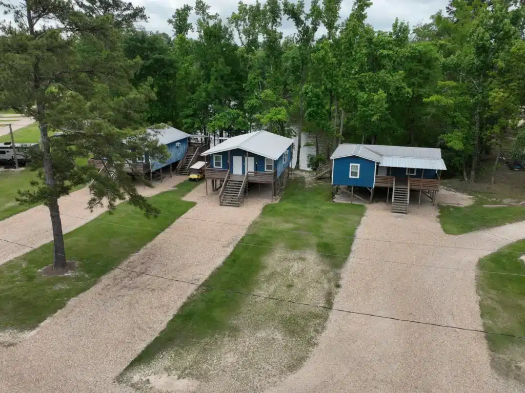 Aerial view of three blue elevated cabins with metal roofs and wooden stairs beside gravel drives and wooded lakefront.