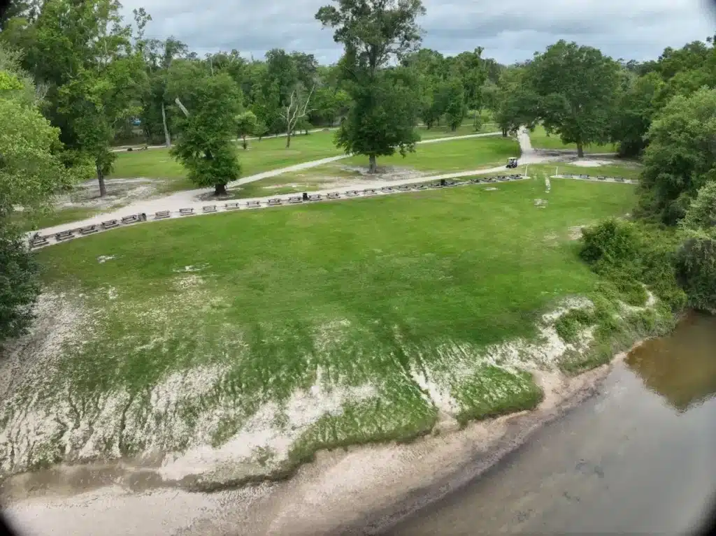 Aerial view of a grassy riverside park with a long row of picnic tables lining a paved path near trees.