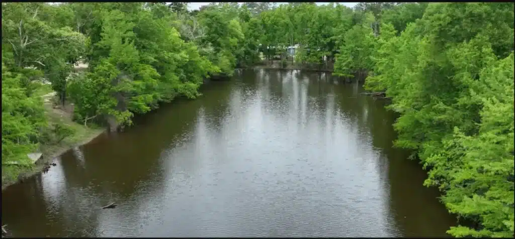 Aerial view of a calm dark river or lake bordered by dense green trees, with a small campground visible in the distance.