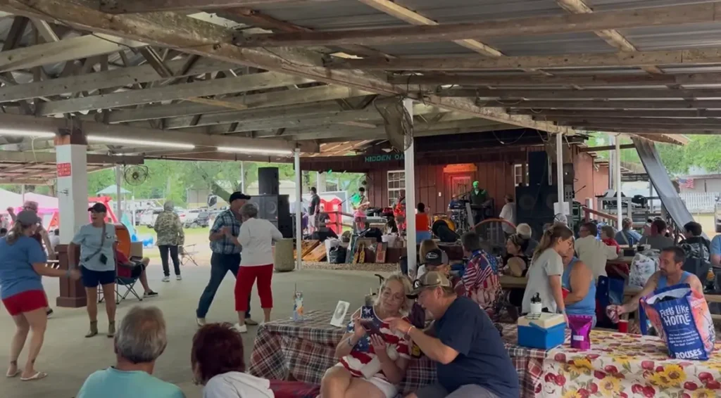 Crowd of people dancing and sitting at picnic tables under a large open-air wooden pavilion with live band playing.