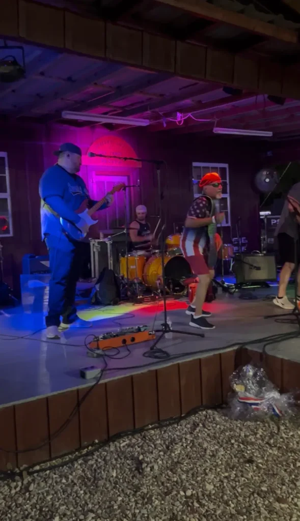 Live band performs on a covered outdoor stage with guitarist, drummer and singer under pink and blue stage lighting.