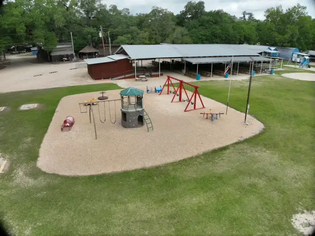 Aerial view of a gravel playground with swings, climbing tower and spring riders beside a large covered pavilion.